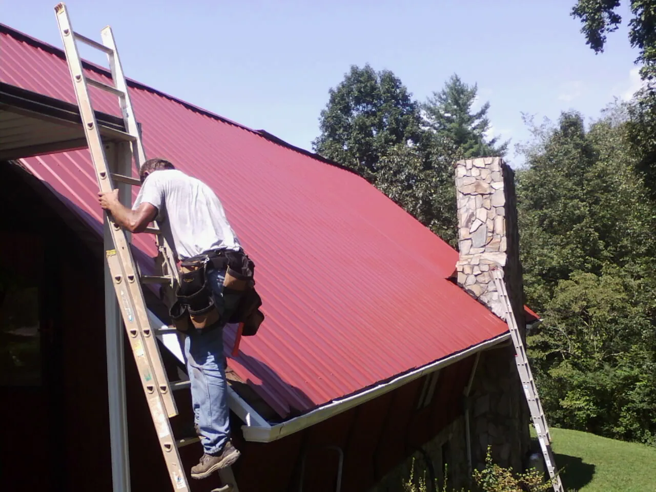 Roofer on a ladder beside a red metal roof and stone chimney — Soffit & Fascia Repair by Roofing Brothers in Round Top